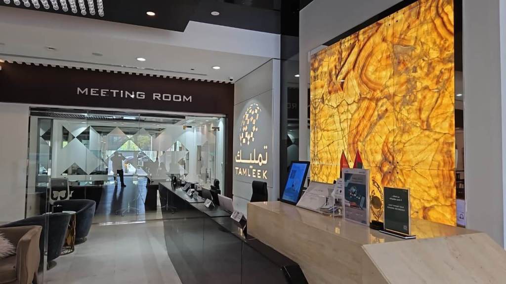Interior view of a modern office lobby with a reception desk and an illuminated wall feature. A sign reads 'MEETING ROOM' and the company's logo is displayed prominently.