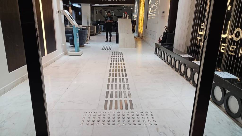 Interior view of a modern lobby featuring a sleek marble floor, metal drainage strips, a meeting room sign in the background, and a person standing near a reception area.