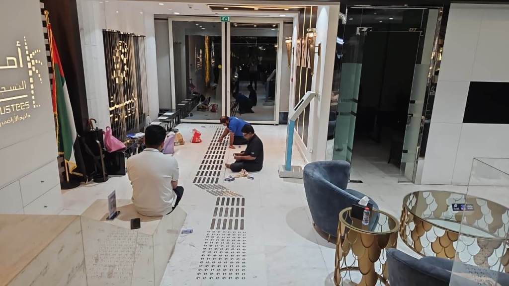 An interior view of a modern, well-lit reception area with a stylish design. People are seated on the floor, engaged in various activities. There are decorative chairs and tables in the foreground, and the walls feature elegant signage in Arabic.