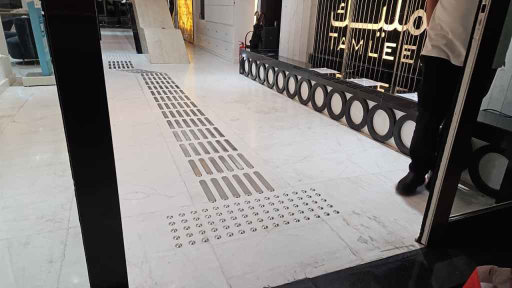 Interior view of a modern space showing a decorative tiled floor with a pattern of narrow and wide slots, leading towards a reception area with a sign that reads 'TAMLEEL'.