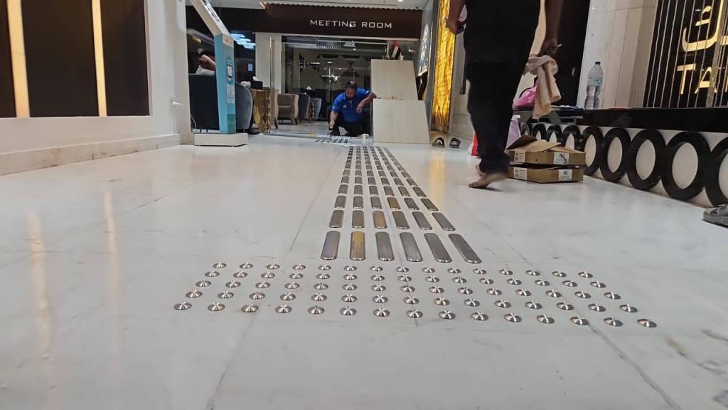 A view of a corridor with a tactile surface on the floor, featuring metal strips and domes, while people walk nearby.