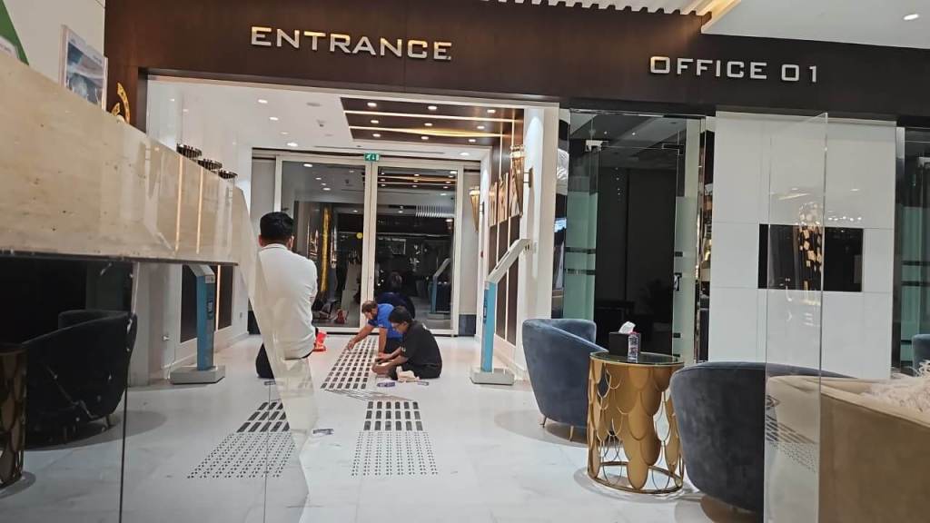 Interior view of a modern office entrance, featuring a sign that reads 'ENTRANCE' and 'OFFICE 01'. People are seen sitting on the floor and near stylish chairs, with a contemporary decor and lighting.