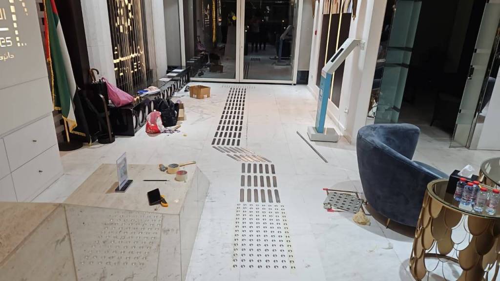 Interior view of a modern establishment showing a marble floor with a decorative strip design, a blue chair, and various items scattered around, including bags, boxes, and construction materials.