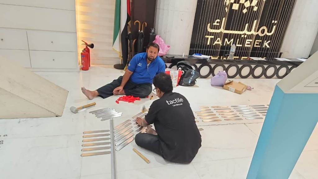 Two men working on the floor of a spacious lobby, assembling tactile strips. One man is dressed in a blue shirt while the other is wearing a black 'tactile' shirt. Nearby, there are items including a fire extinguisher and umbrellas, with a sign reading 'TAMLEEK' in the background.