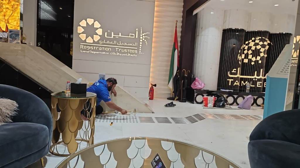 A worker in a blue shirt is applying tiles on the floor in a modern office space, featuring a registration desk area with signage in Arabic and English. In the background, there is a UAE flag and various items are placed around the room.