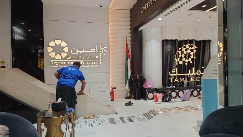 A worker wearing a blue shirt is adjusting a ramp in a modern office lobby. The background features the logos of 'Tamleek' and 'Registrars of Trustees,' with a UAE flag and various items scattered on the floor.