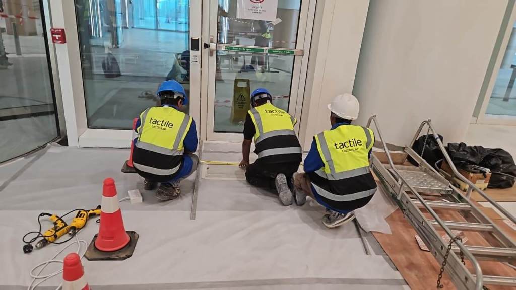 Three workers in safety vests and helmets are installing equipment near a door, with construction materials and tools nearby.