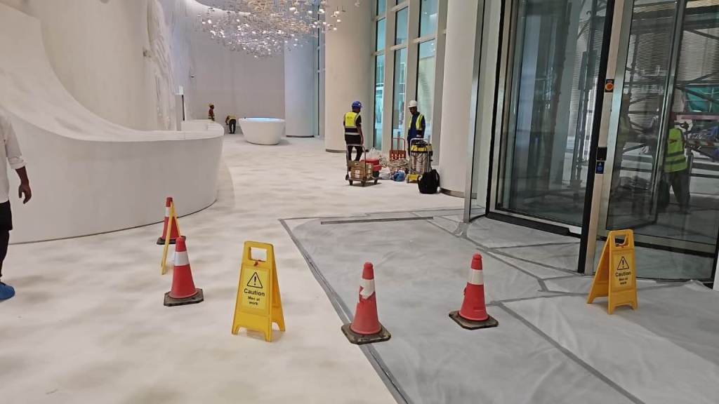 Interior of a building under construction, featuring a white floor, caution cones, and construction workers in safety gear.