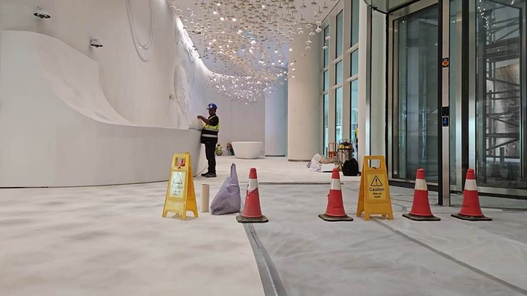 A partially completed interior space featuring a smooth white wall, scattered construction materials, and a worker in a safety vest. Caution signs and traffic cones are arranged to indicate a work zone. Decorative lighting hangs from the ceiling.