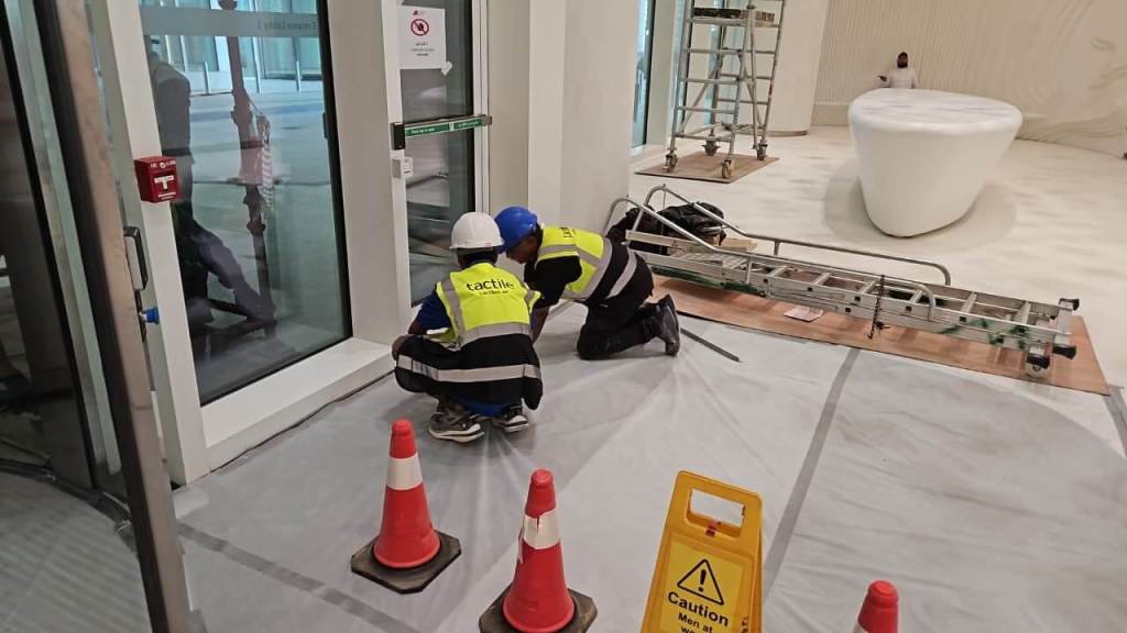 Two construction workers in hard hats and safety vests kneeling near a glass door, working on installation, with safety cones and a caution sign nearby.