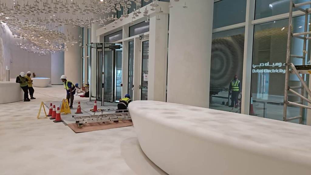 Interior view of a modern lobby under construction, featuring a white curved reception desk, workers in safety vests and hard hats, and construction tools. Decorative elements hang overhead.