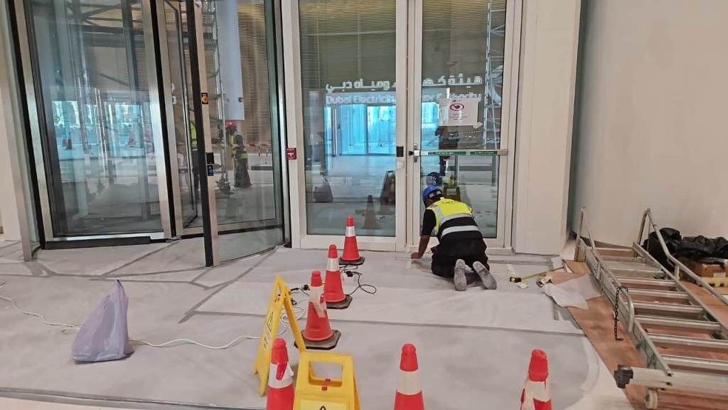 A construction worker kneeling on the floor near glass doors, surrounded by traffic cones and caution signs. The area is under renovation with visible tools and materials.
