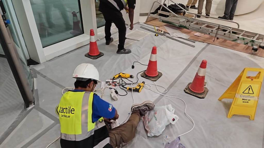 A construction worker in a yellow safety vest and helmet is seated on the floor working with electrical tools. Nearby, there are traffic cones, a caution sign indicating 'Men at work', and a ladder. The floor is covered with protective sheeting.