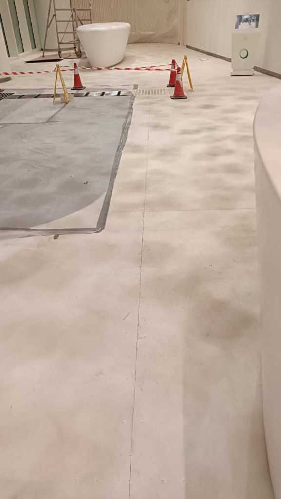 Interior view of a partially finished room showing a light-colored floor, construction barriers, and a white decorative tub. A ladder is visible in the background.