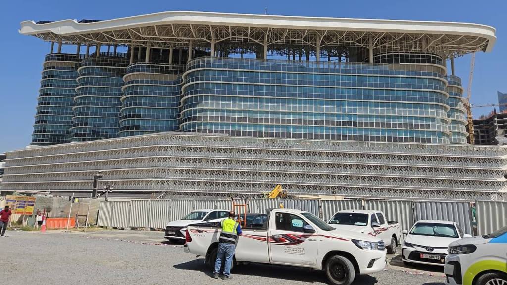 A modern multi-story building under construction with a distinctive architectural design, featuring large glass windows and tiered levels. Several vehicles are parked in the foreground, with construction barriers visible nearby.