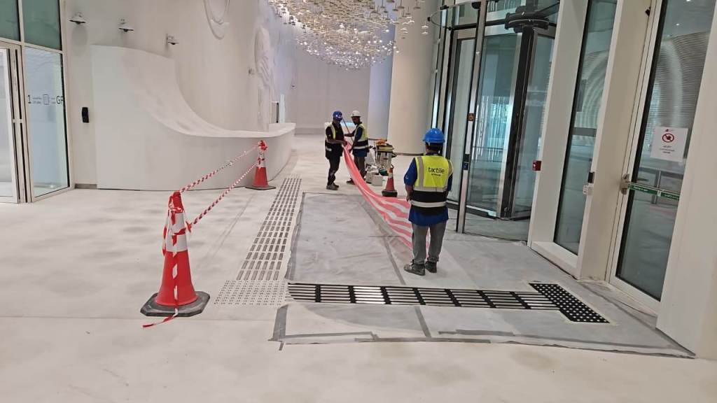 Construction workers in safety gear are working on a polished, unfinished floor with safety cones and barriers. The area features modern architecture with white walls and a decorative lighting installation overhead.