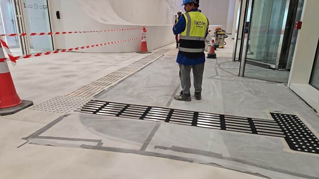 A construction worker standing on a partially finished floor with tactile paving and safety cones in the background.