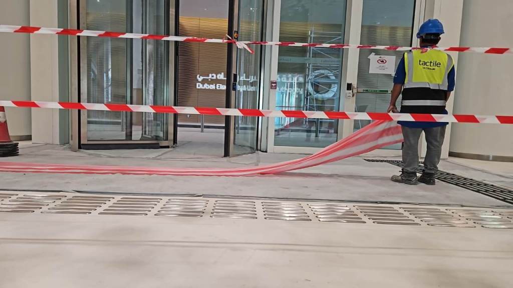 Construction worker in a blue helmet and reflective vest stands near a closed entrance, with red and white caution tape surrounding the area.