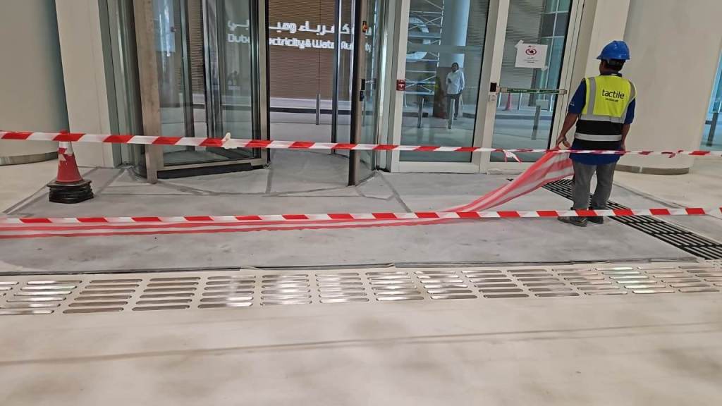 A construction worker in a blue helmet and a reflective vest is standing near a glass door entrance that is cordoned off with red and white caution tape. A traffic cone is positioned nearby, and the floor appears to be under renovation.