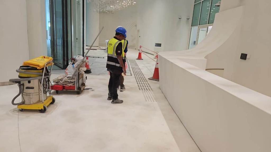 Two construction workers wearing safety vests and helmets, standing in a partially finished interior space, with cleaning equipment and traffic cones nearby.