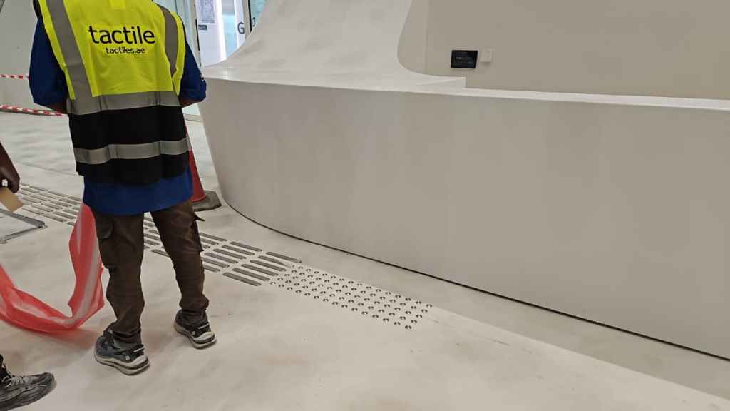 A construction worker in a neon yellow vest stands near a white curved wall in an unfinished interior space, with floor markings visible and a red safety tape in the background.