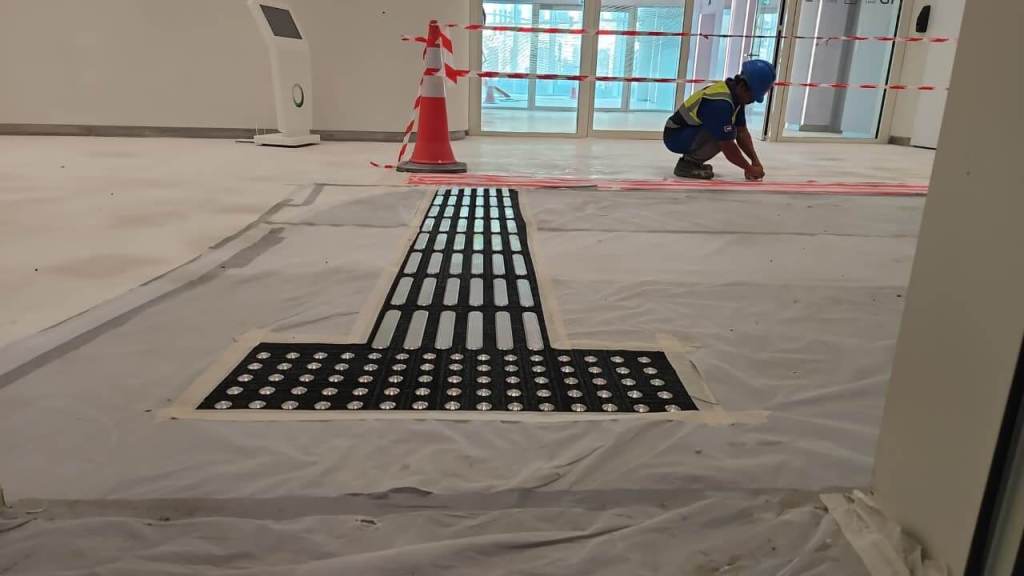 A construction worker wearing a blue helmet and reflective vest is kneeling on a partially finished floor. A textured black mat with circular patterns is laid out in the foreground, while safety cones and caution tape indicate restricted areas in the background.