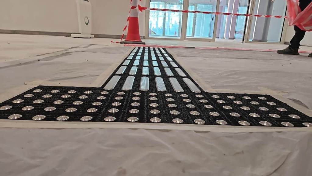 A close-up view of a tactile ground surface indicator with raised patterns, set on a construction site with a plastic floor covering. Safety cones and barrier tape are in the background.