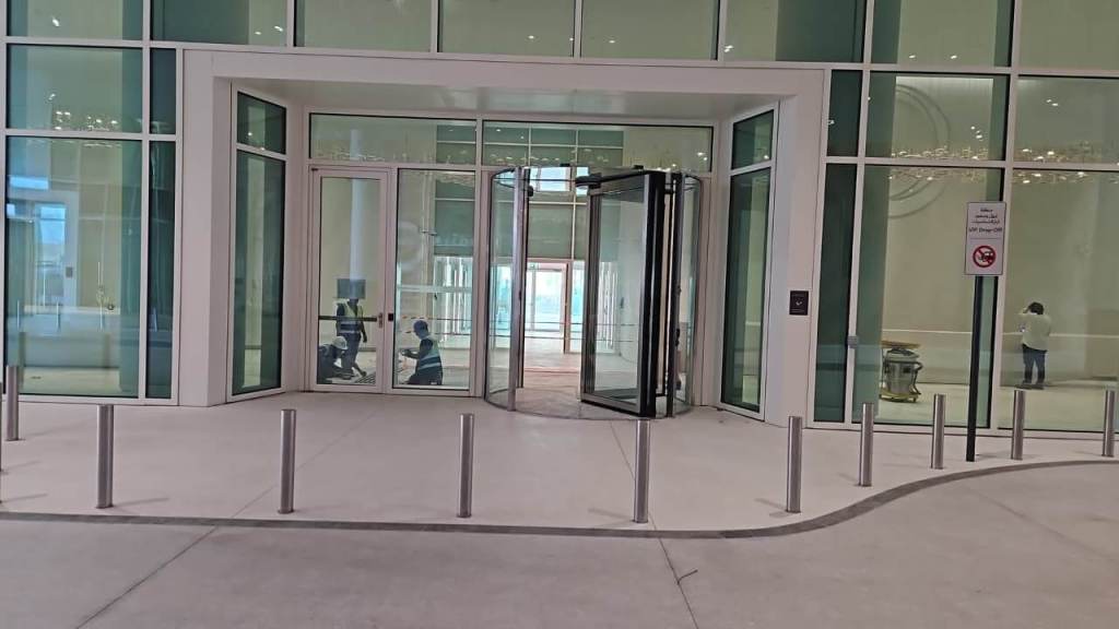 Entrance of a modern building featuring glass doors and a revolving door, with an open interior visible. A sign indicating no smoking is posted nearby.