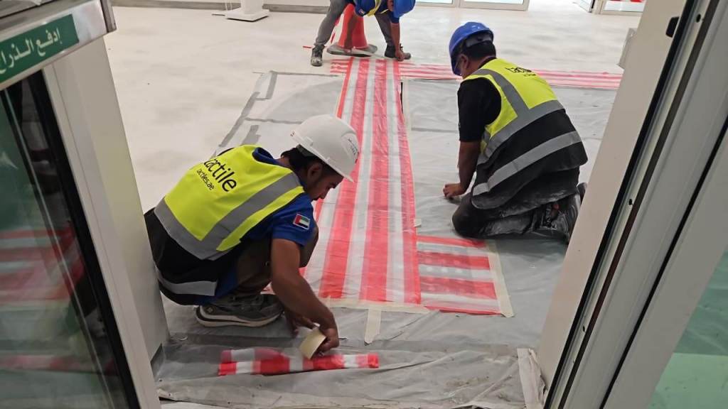 Two construction workers applying red and white striped tape on a floor, with one kneeling and the other sitting. Both are wearing safety vests and helmets.