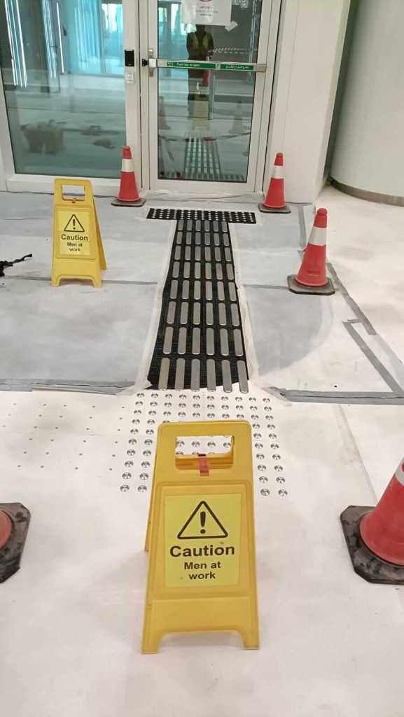 Construction area with caution signs, cones, and a guiding pathway leading to an entrance.
