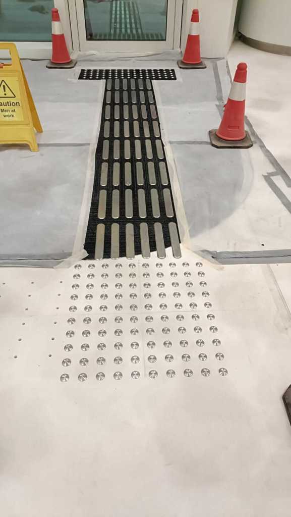 A walkway with tactile paving leading to a doorway, surrounded by traffic cones and a caution sign indicating ongoing work.