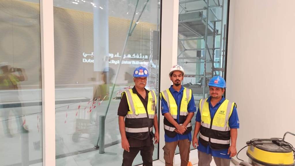 Three construction workers wearing safety vests and hard hats, posed in front of large glass windows in a building under construction.