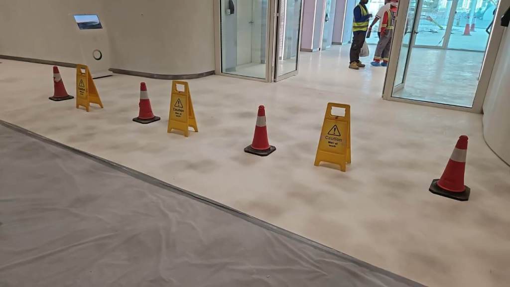 Interior view of a construction site with yellow caution signs and traffic cones on a white floor. Workers are present in the background, and glass doors lead to other areas.