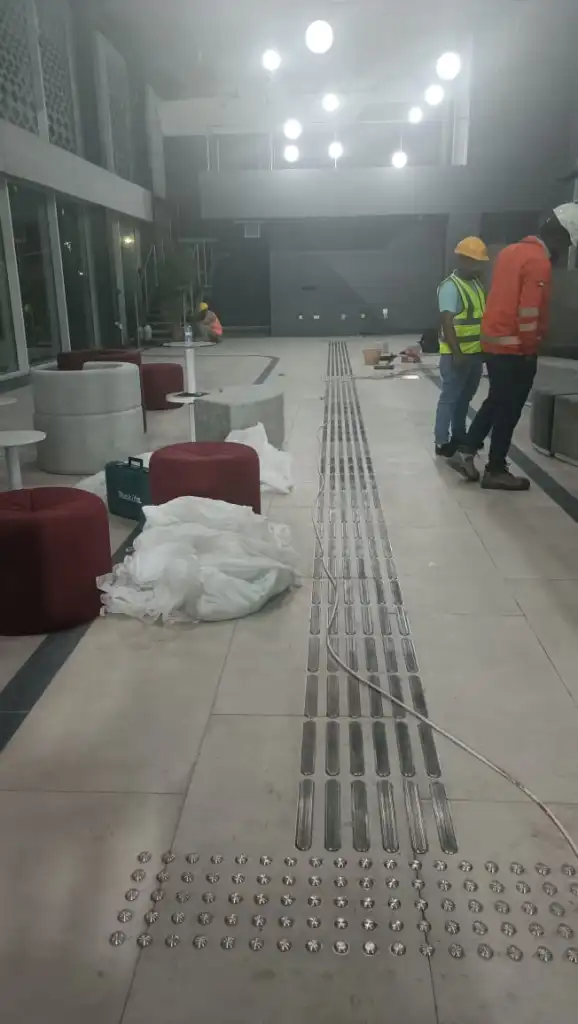 An interior construction site with two workers, one in a yellow hard hat and another in a red jacket, discussing in a spacious room. The flooring features metallic strips and circular fixtures, with unfinished furniture and construction materials visible.