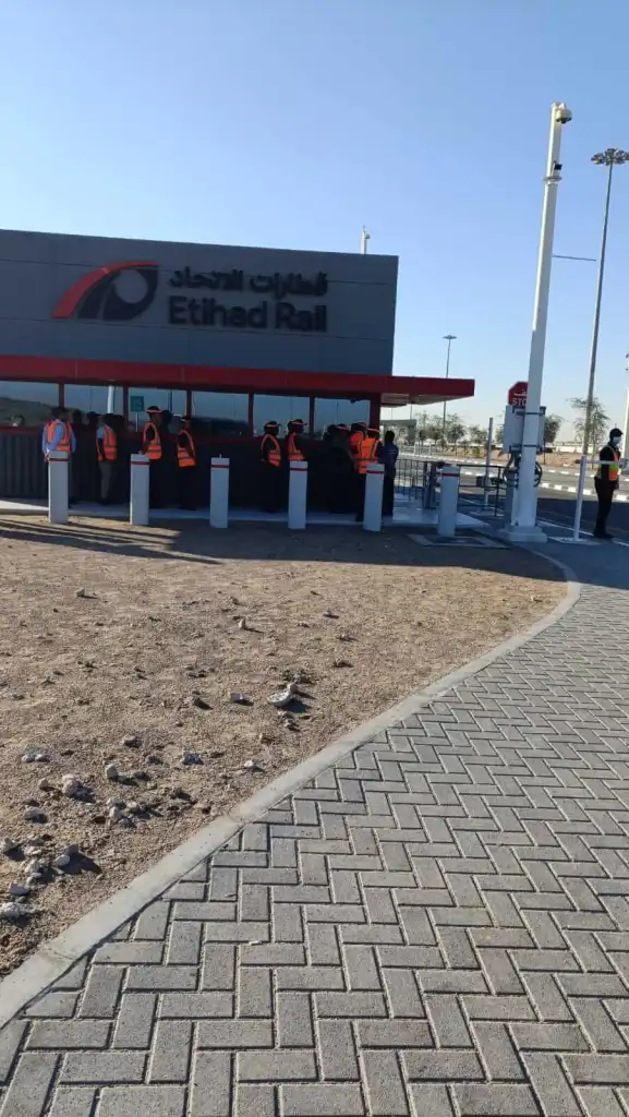 A group of people in safety vests gathered at the Etihad Rail station entrance, with the station building and clear blue sky in the background.