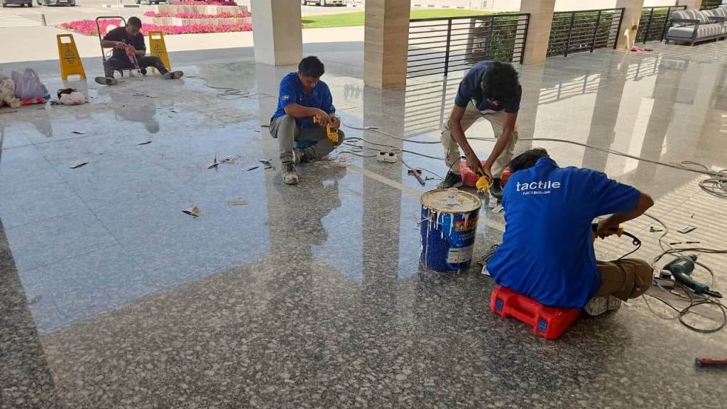 Workers performing floor maintenance on a shiny tiled surface, using tools and equipment. Warning signs for wet floor are visible.