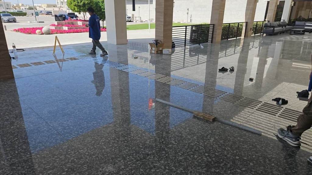 A worker walking on a polished stone floor inside a building, with tools and safety signs visible. The area features large windows and outdoor greenery.