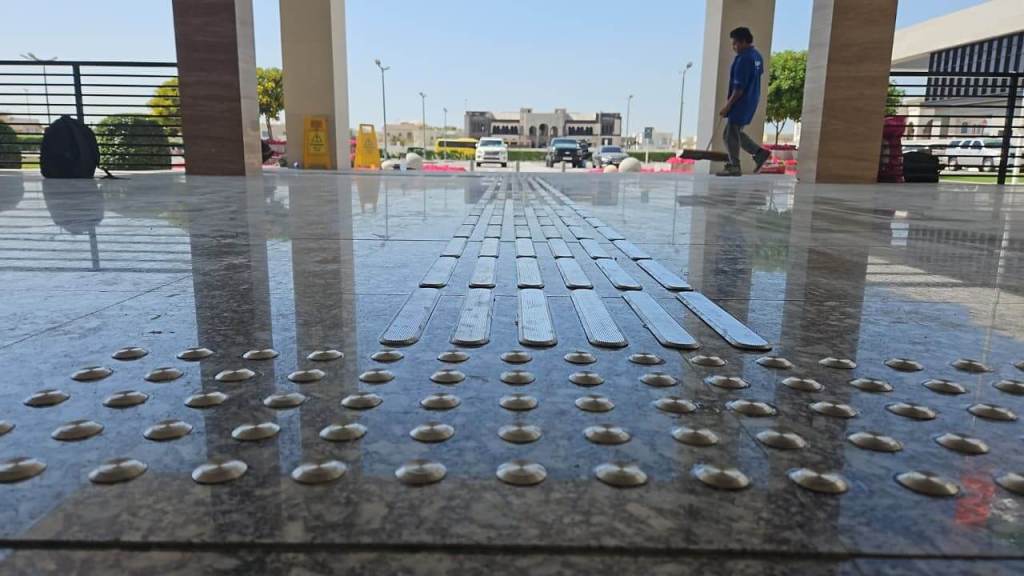 A close-up view of a textured floor featuring tactile paving with dot patterns and ridges, designed for accessibility, alongside a walkway where a person is walking in the background.