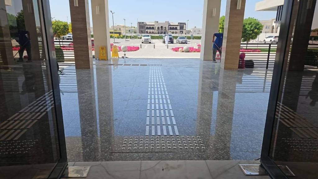 View of the entrance to a modern building with polished flooring, showing a pathway with tactile strips and a sunny outdoor area with parked cars and landscaped greenery.