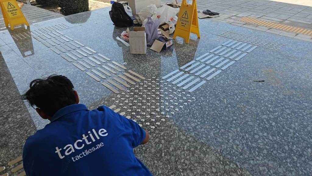 A worker kneels on a polished surface, applying tactile paving strips and tactile indicators. Surrounding materials and caution signs indicate ongoing construction or maintenance work.