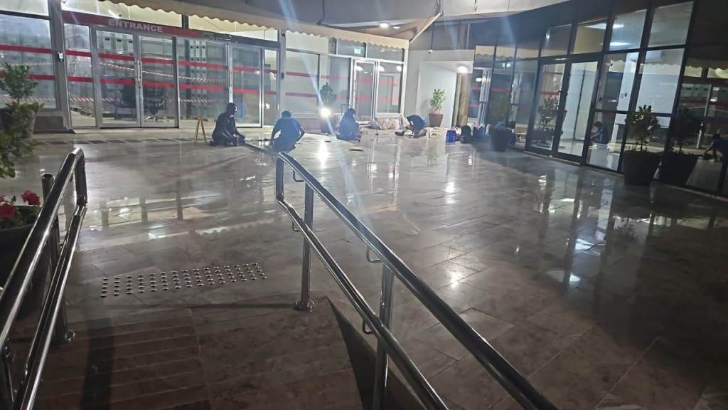 A dimly lit entrance area with polished flooring, a few people sitting on the ground, and potted plants near large glass doors.