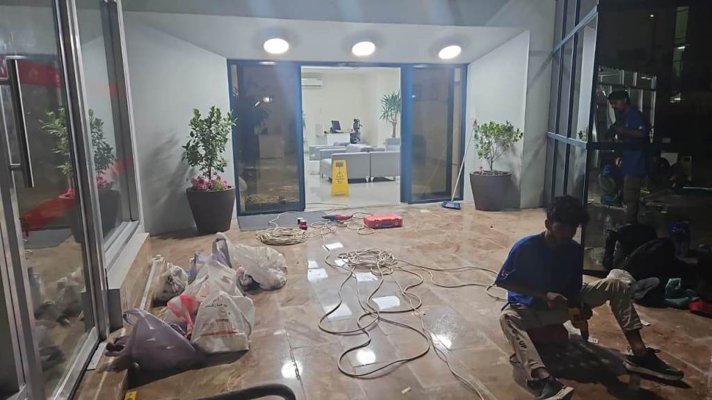 Interior of a partially constructed office space with a polished floor, featuring potted plants and construction materials scattered around. Two workers are seen: one sitting on the floor using a handheld tool, and another working near the entrance.