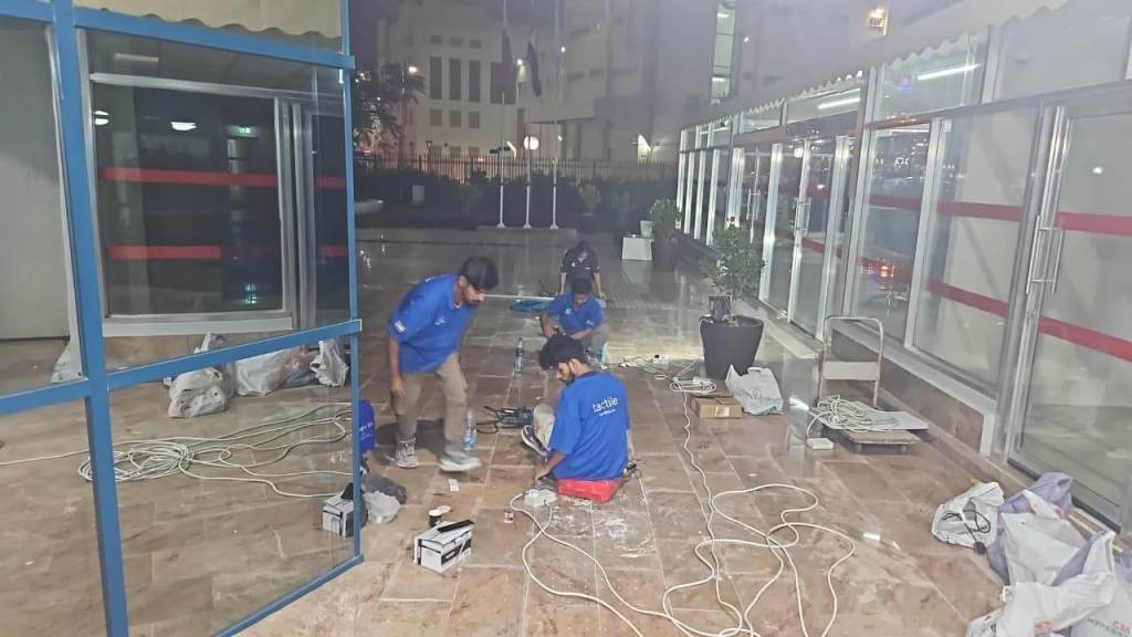 Three workers in blue shirts are setting up equipment and cleaning the floor in an outdoor area at night. Tools and materials are scattered around, and a potted plant is visible in the background.