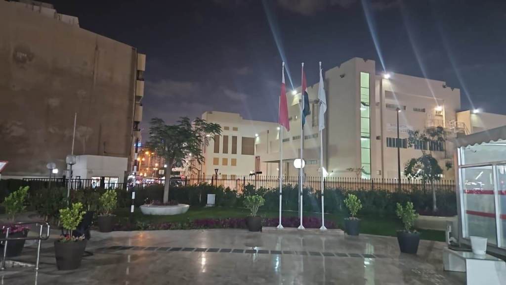Nighttime view of a modern building with flags and landscaped greenery in the foreground.