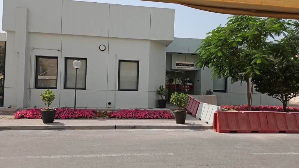 Exterior view of a modern building with large windows, surrounded by colorful pink flower beds and potted plants, with a barrier in the foreground.