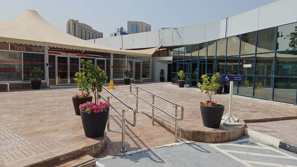 Entrance to a building with a ramp and potted plants, featuring a large awning and glass windows.