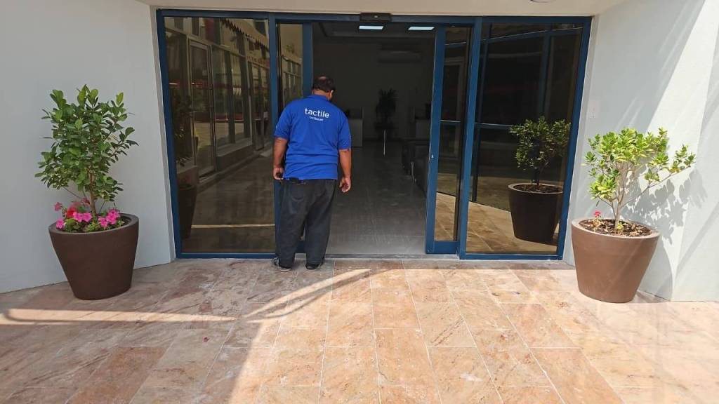 A person wearing a blue shirt is standing in front of an entrance with glass doors, looking inside. There are potted plants on either side of the entrance, with one pot containing pink flowers.