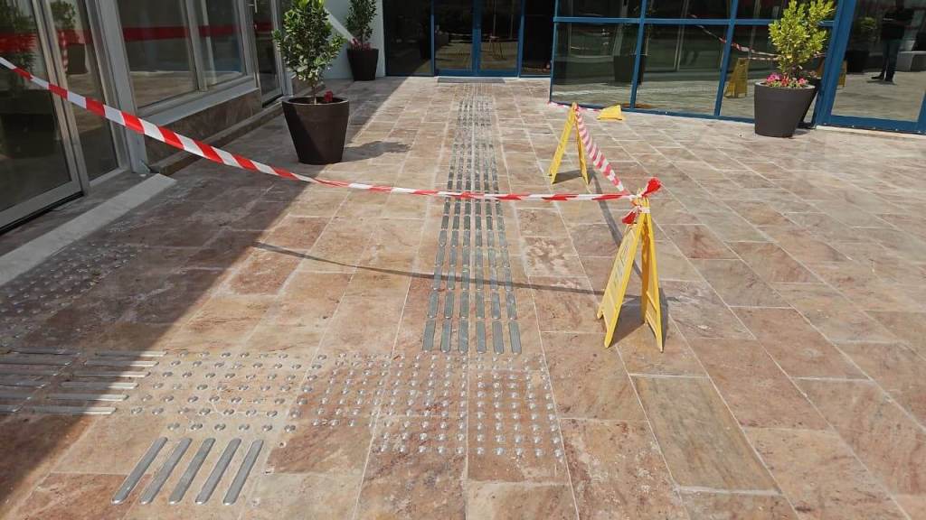 An outdoor area with tiled flooring, marked off by caution tape and warning signs. There are potted plants nearby and visible tactile paving strips on the ground.