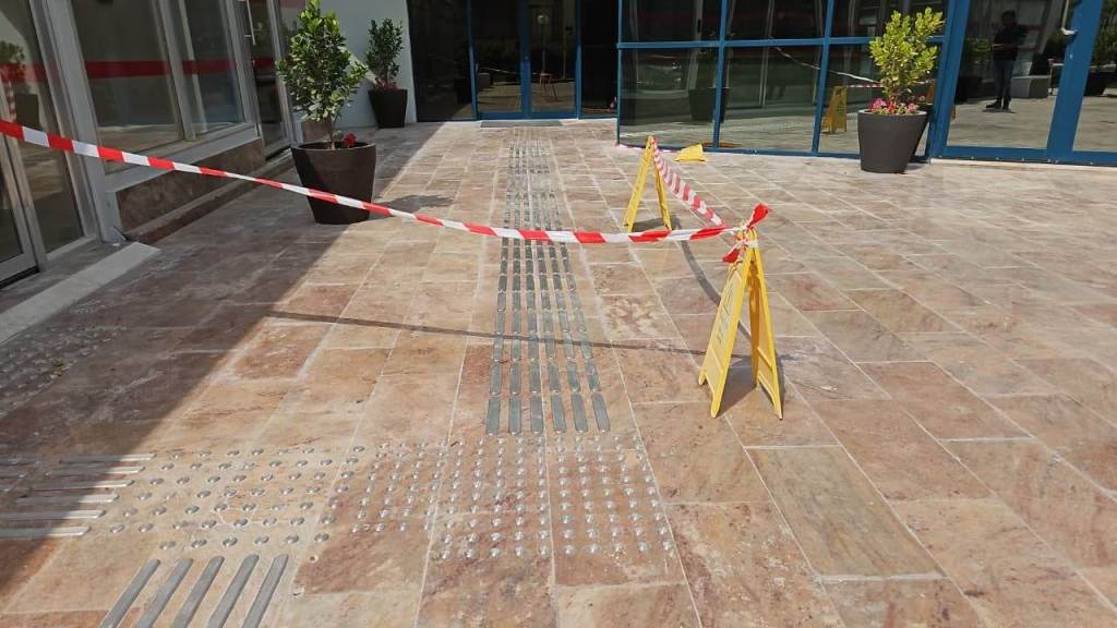 A view of a tiled outdoor area with caution tape and warning signs indicating restricted access, featuring decorative potted plants and a glass entrance.