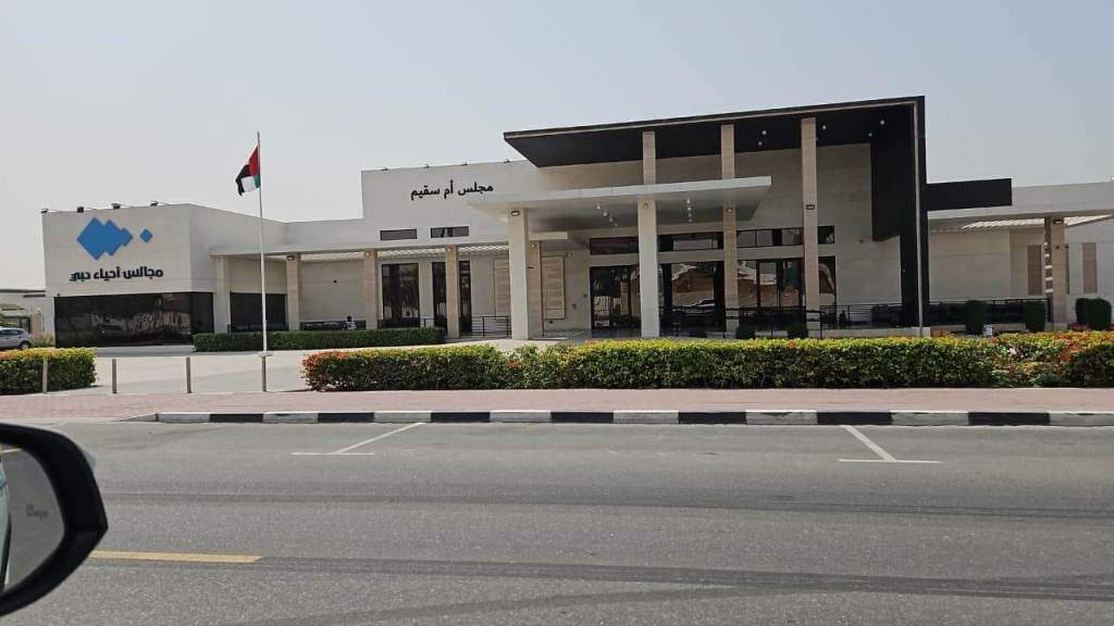Exterior view of a modern building with a black and white facade, featuring large glass doors and landscaped greenery in front. The UAE flag is displayed on a flagpole.